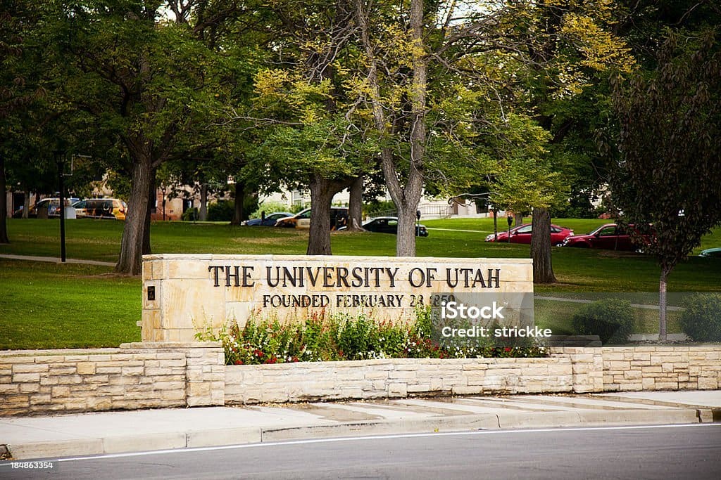 Large stone engraved with 'University of Utah' surrounded by scenic landscapes and clear skies.